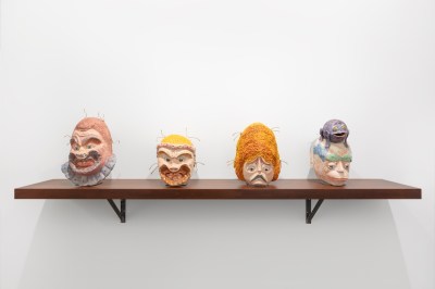 Four sculpted heads set on a wooden shelf. Two of the heads appear to smile really big, the third appears to frown, and the fourth looks like a monkey.