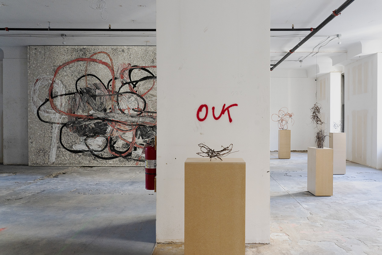 In the background, a garbled scribbly mosaic. In the foreground, a column is spray painted with the word "out." Throughout the space, garbled wire tumbleweeds are perched on wooden pedestals.