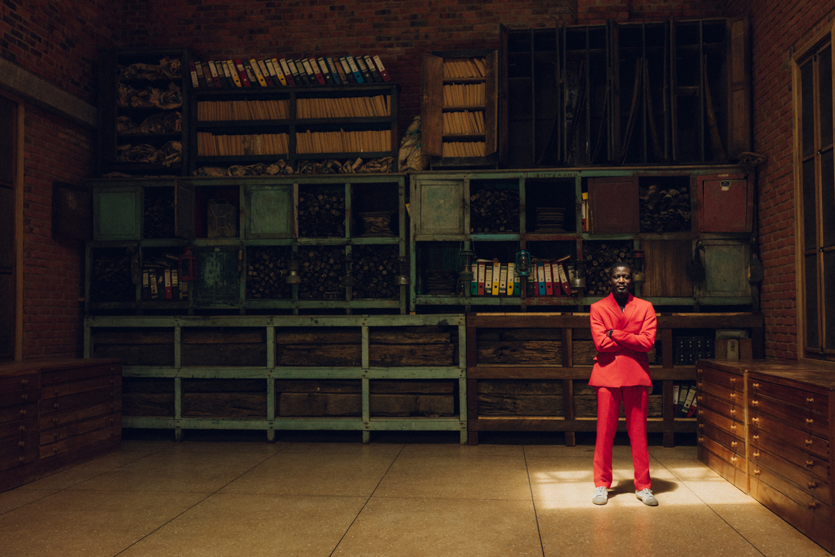 Portrait of Ibrahim Mahama, a Black man in a red suit stands in a square of light in front of an archive.