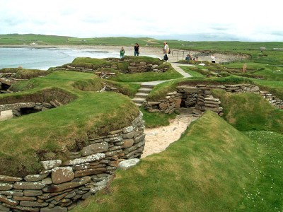 A few people standing around the remains of the Skara Brae village in Scotland, with green grass-covered mounts and stone steps