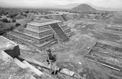 black and white photo of Teotihuacan showing stone archaeological structures and a person with binoculars climbing the ancient steps in the foreground