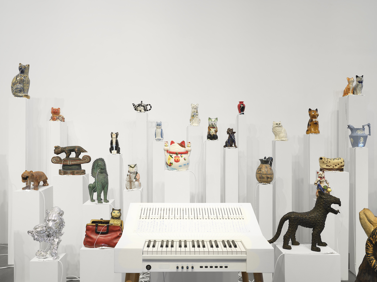 A series of cat vessels and figurines on pedestals at various heights in front of a white keyboard that triggers sounds within each.