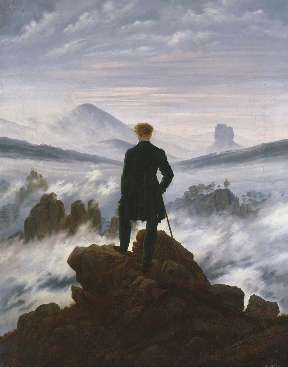 A man stands on a cliff with his back to the viewer, overlooking mountaintops that poke out of misty clouds.