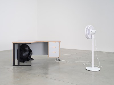 A sculpture of a gorilla crouched beneath a desk. A plugged-in fan is pointed at the structure.