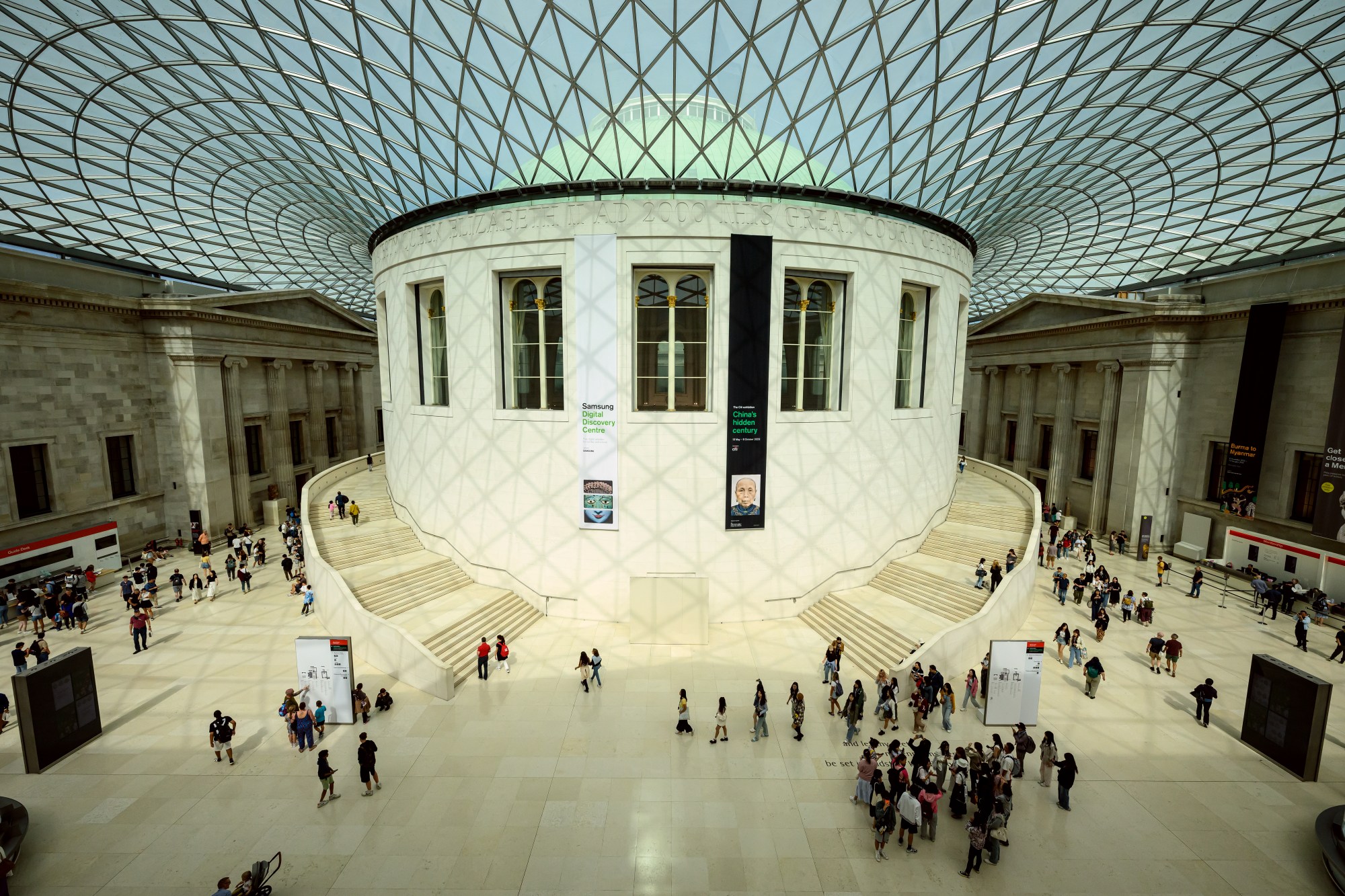 In a color photo, a futuristic translucent ceiling hangs over a large atrium with classical architecture. It is filled with small people.