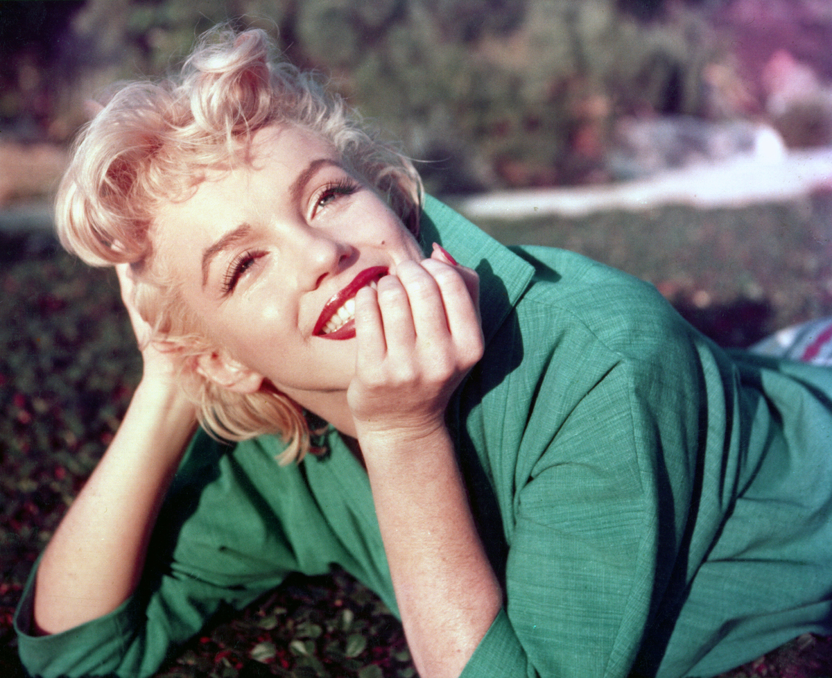 A white woman smiling and lying down on grass, propping her head up with her arms.