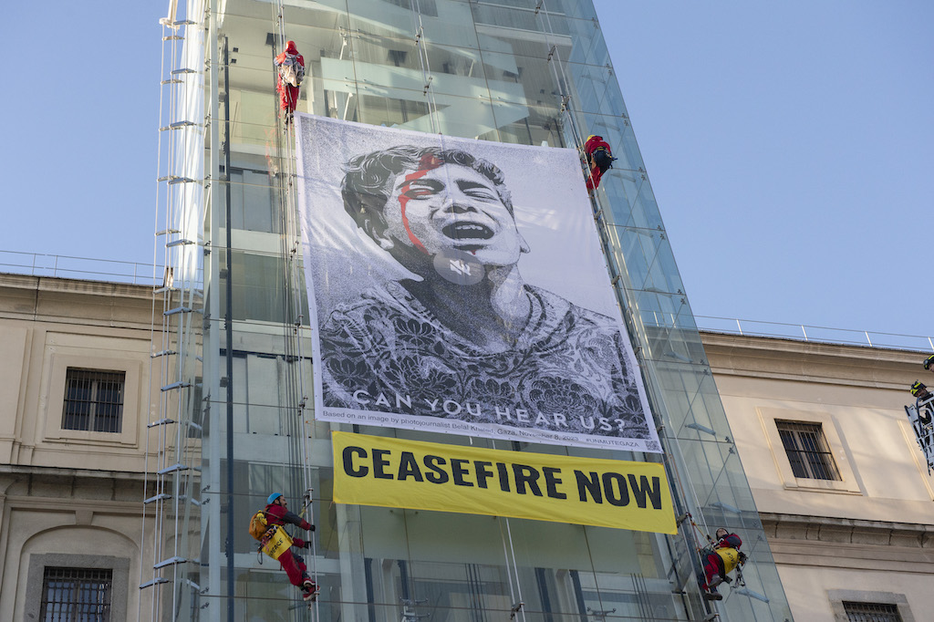 A group of people mounting a scaffold and mounting an image of a boy crying with blood running down his head. Printed beneath the image are the words 'CAN YOU HEAR US?' A yellow banner reading 'CEASEFIRE NOW' is also attached.