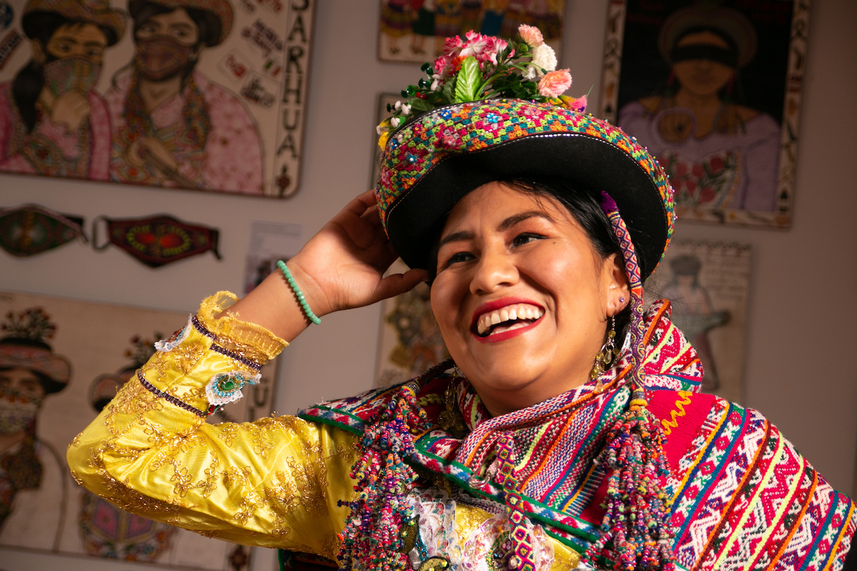 A smiling woman in multicolored woven clothing in front of several paintings.