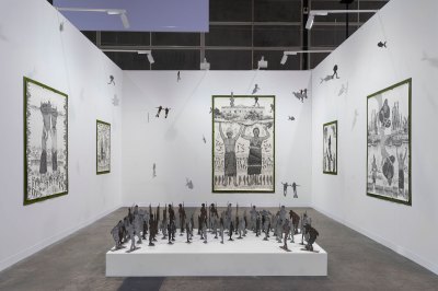 View of an art fair booth showing various drawings showing people and an installation of cutout people on a plinth at front.