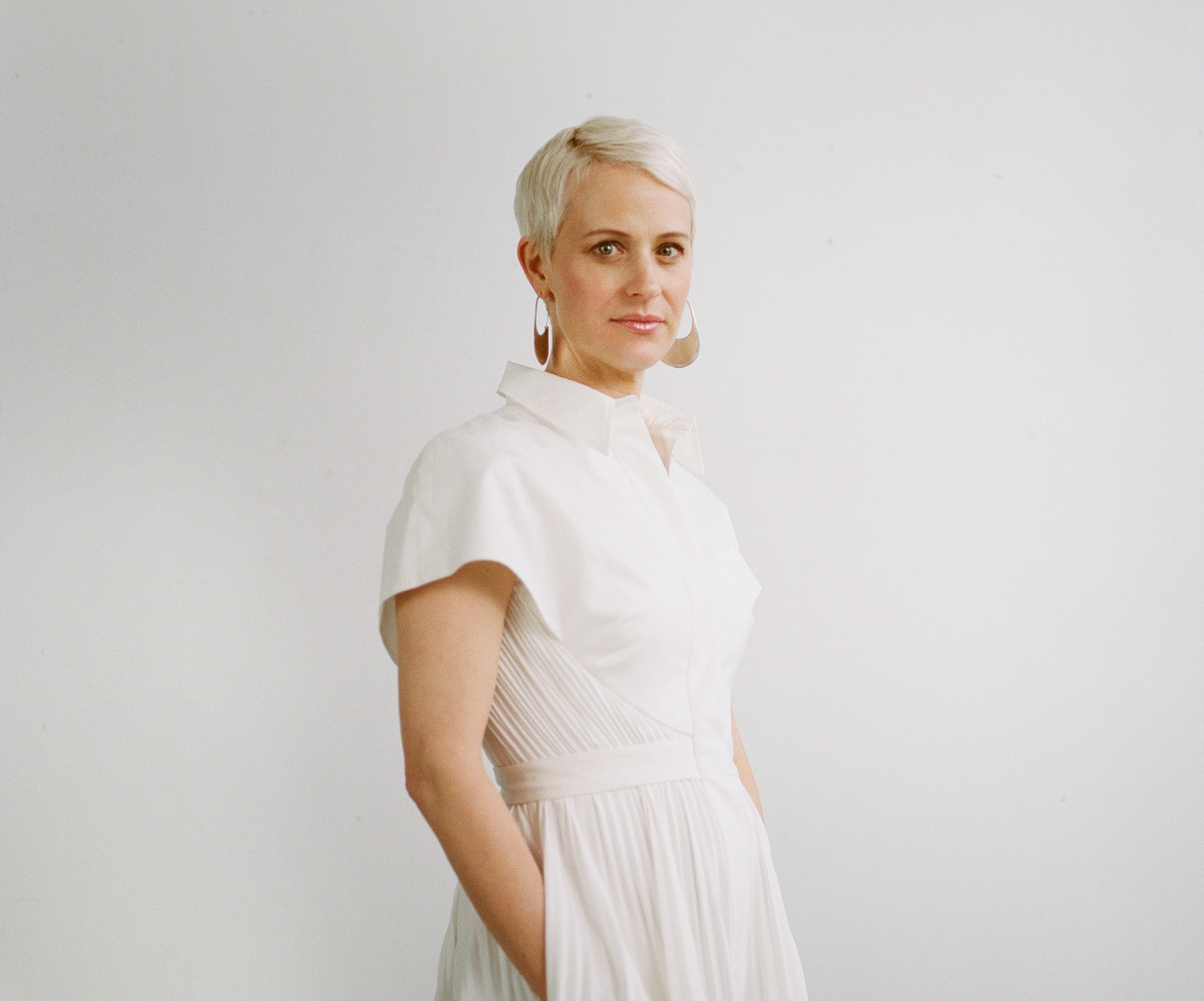 A woman with platinum blonde hair poses in front of a white background. She wears a white dress and half-crescent metal earrings.