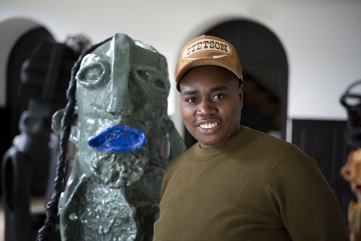 A Black woman in a baseball cap standing beside a clay sculpture with gaping blue lips.