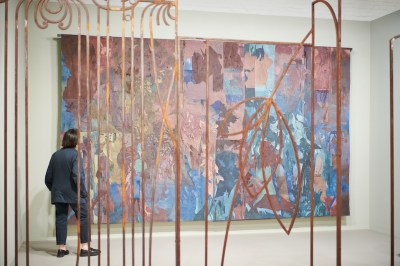 A person seen from behind staring at a large, abstract textile piece in shades of brown and blue. A metal, gate-like structure can be seen in front.