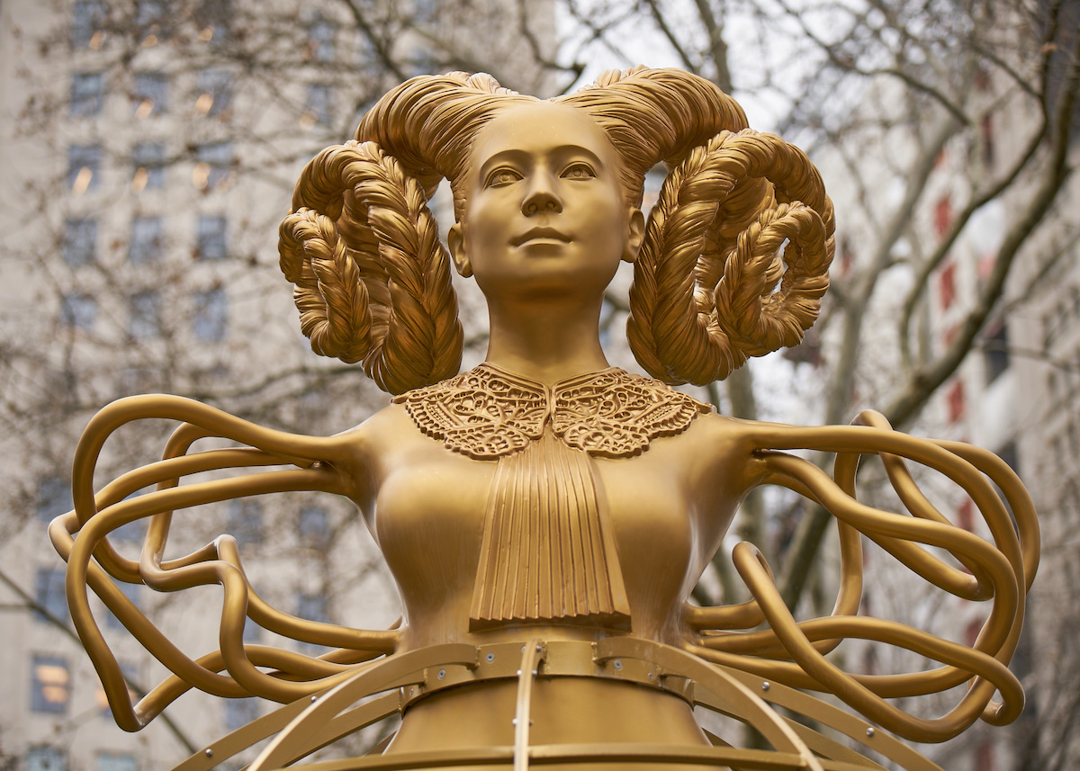 A gold-toned sculpture of a woman with spiraling braids of hair. In place of her arms, she has winding, intersecting abstract forms.